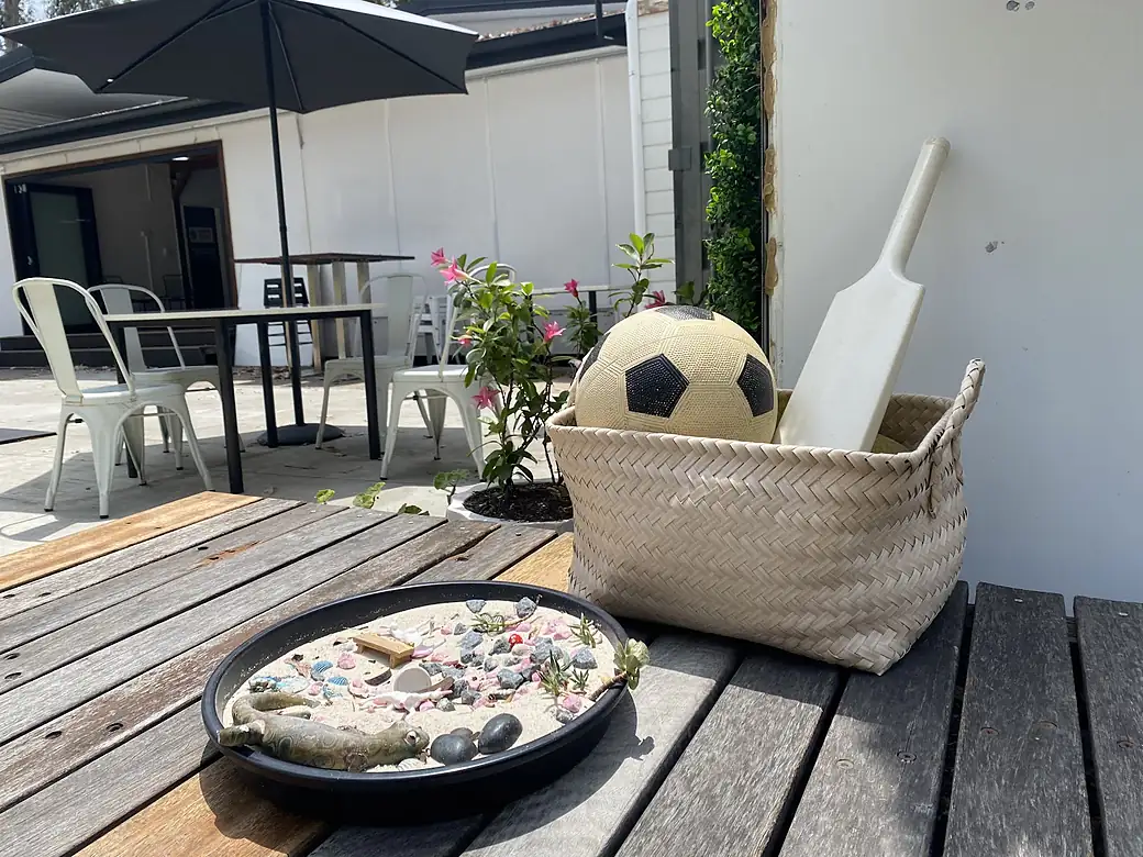 A sand tray, a football and a bat in a basket on the timber deck, with tables and an umbrella behind